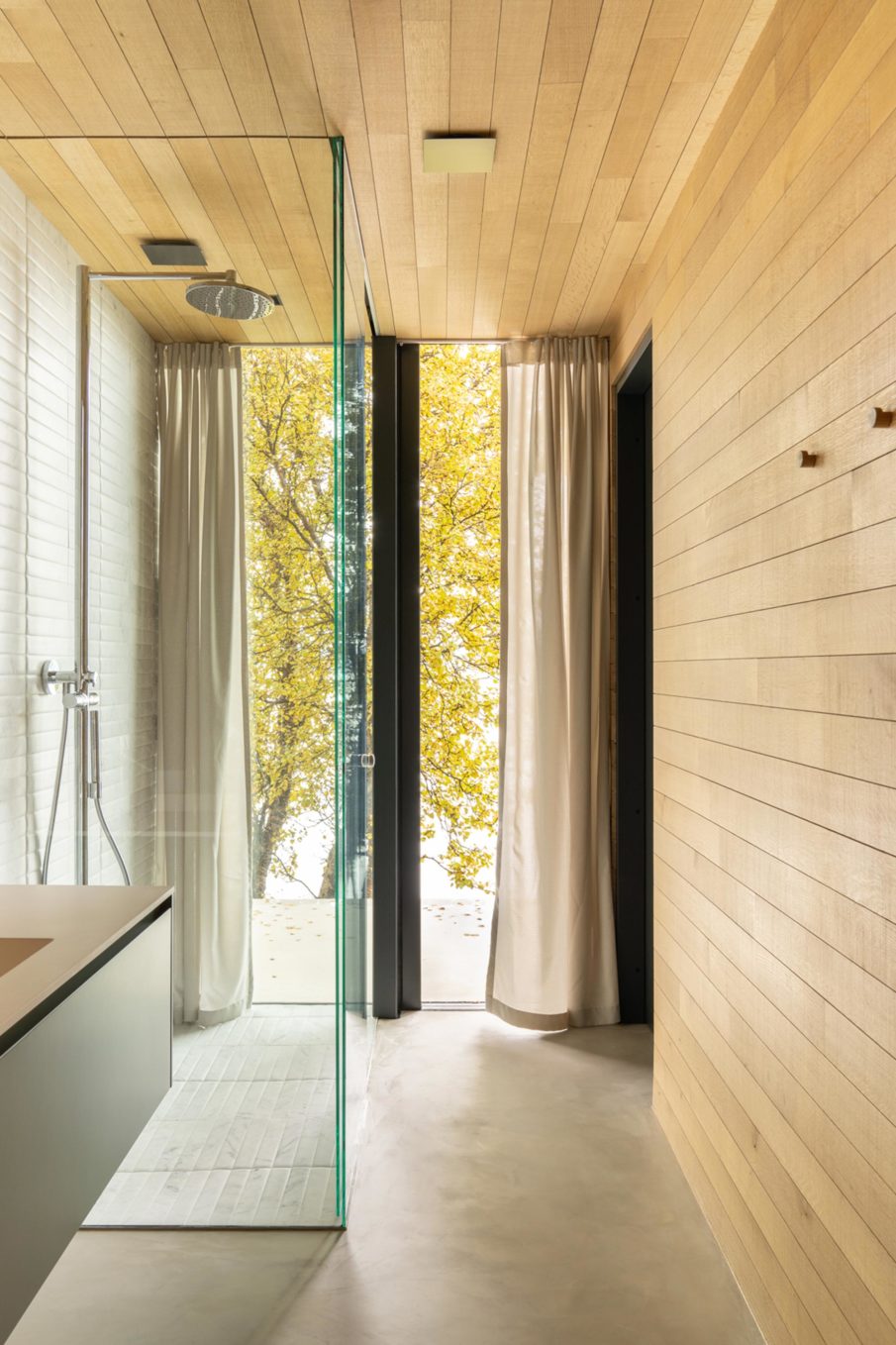 A part of a bathroom with a shower and large windows with curtains that can be drawn.
