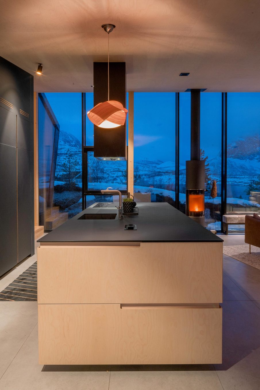Kitchen island with a fireplace in the background, dusk outside behind the large windows.